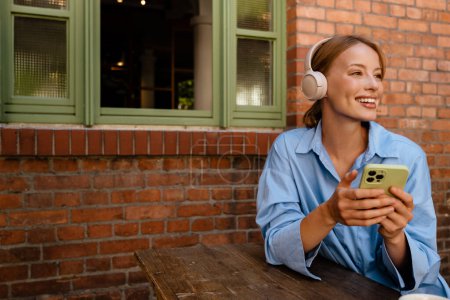Téléchargez les photos : Ginger white woman in headphones using cellphone while sitting at cafe indoors - en image libre de droit