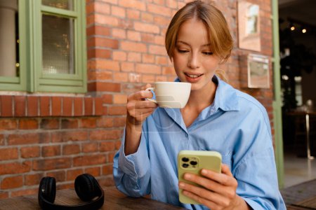 Téléchargez les photos : White young woman using mobile phone while drinking coffee in cafe outdoors - en image libre de droit