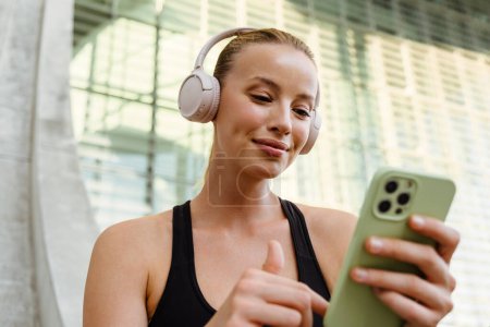 Photo for White young woman in headphones using mobile phone after yoga practice outdoors - Royalty Free Image