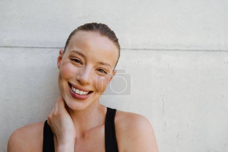Téléchargez les photos : Jeune femme blanche en vêtements de sport souriant à la caméra tout en posant par mur à l'extérieur - en image libre de droit