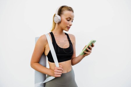 Téléchargez les photos : Jeune femme blanche dans les écouteurs en utilisant le téléphone mobile après la pratique du yoga en plein air - en image libre de droit