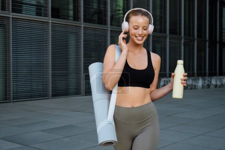 Photo for White young woman wearing headphones walking with water bottle after workout outdoors - Royalty Free Image