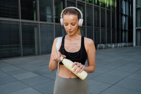 Foto de Mujer joven blanca con auriculares caminando con botella de agua después del entrenamiento al aire libre - Imagen libre de derechos