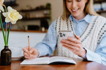 Foto de Mujer blanca rubia usando el teléfono móvil mientras escribe notas en el café en el interior - Imagen libre de derechos