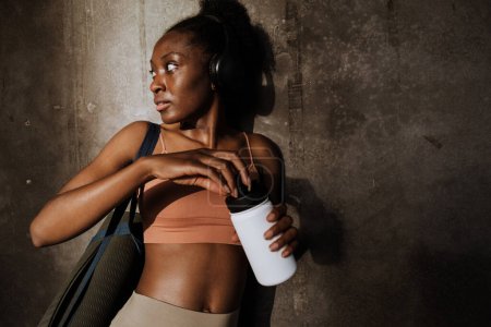 Photo for Young beautiful sporty calm african woman in headphones with yoga mat opening bottle of water and looking aside , while standing near grey wall - Royalty Free Image