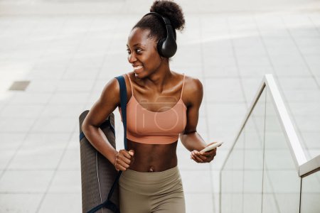 Téléchargez les photos : Young beautiful smiling fit african woman in headphones with phone and yoga mat looking aside, while walking stairs outdoors - en image libre de droit
