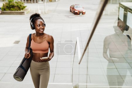 Téléchargez les photos : Jeune belle femme africaine souriante en casque avec téléphone et tapis de yoga regardant de côté, tout en marchant les escaliers à l'extérieur - en image libre de droit