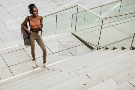 Foto de Young beautiful smiling fit african woman in headphones with bottle and yoga mat looking aside, while walking stairs outdoors - Imagen libre de derechos