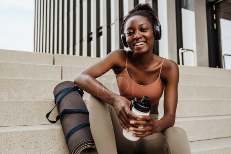 Téléchargez les photos : Young beautiful smiling happy fit african woman in headphones with bootle and yoga mat looking at camera , while sitting on stairs outdoors - en image libre de droit