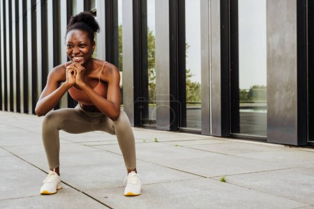 Photo for Young beautiful sporty smiling african woman crouches with folded hands and looking aside, while standing outdoors near modern building - Royalty Free Image