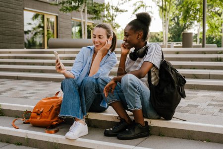 Téléchargez les photos : Les jeunes filles multinationales parlent et utilisent le téléphone portable ensemble tout en étant assis sur les escaliers à l'extérieur - en image libre de droit