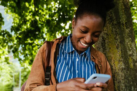 Foto de Retrato de una joven hermosa africana sonriente mujer feliz mirando el teléfono mientras está de pie al aire libre - Imagen libre de derechos