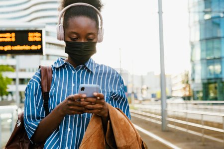 Téléchargez les photos : Jeune femme africaine en casque et masque avec sac à dos regardant son téléphone tout en restant debout à l'extérieur - en image libre de droit