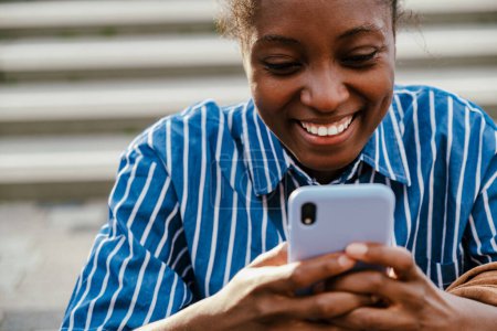 Foto de Portrait of young beautiful african smiling happy woman with phone looking on it while standing outdoors - Imagen libre de derechos