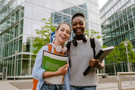 Téléchargez les photos : Jeunes étudiantes multiraciales souriantes et étreintes debout dans la rue de la ville à l'extérieur - en image libre de droit