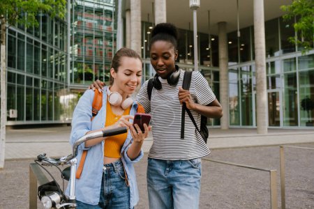 Téléchargez les photos : Young multiracial women smiling and using cellphone while standing at city street - en image libre de droit