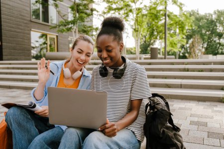 Téléchargez les photos : Young multiracial women doing homework with laptop while sitting on stairs outdoors - en image libre de droit