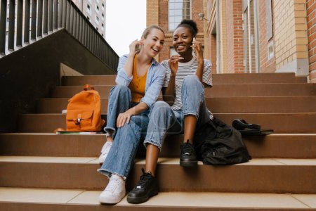 Téléchargez les photos : Les jeunes filles multinationales souriant et utilisant le téléphone portable ensemble tout en étant assis sur les escaliers à l'extérieur - en image libre de droit