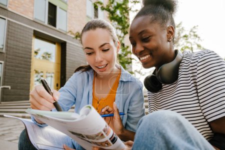Téléchargez les photos : Les jeunes femmes multiraciales souriant et faisant leurs devoirs ensemble tout en étant assis sur l'escalier à l'extérieur - en image libre de droit