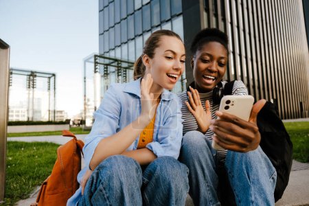 Téléchargez les photos : Jeunes femmes multiraciales agitant les mains et utilisant un téléphone portable alors qu'elles sont assises dans la rue de la ville - en image libre de droit