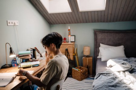 Foto de Asiático adolescente chico usando celular mientras sentado en escritorio y estudiar en casa - Imagen libre de derechos