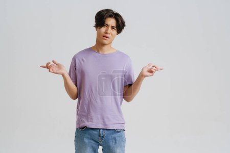 Photo for Perplexed asian man wearing t-shirt pointing fingers at opposite sides isolated over white background - Royalty Free Image