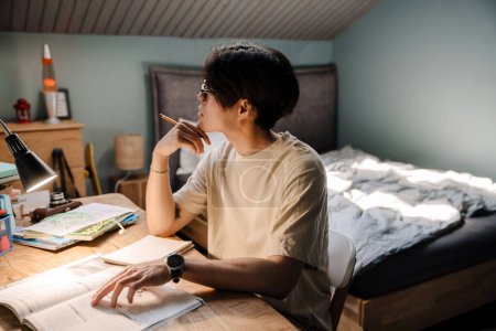 Téléchargez les photos : Serious asian teenage guy studying with book while sitting at desk at home - en image libre de droit