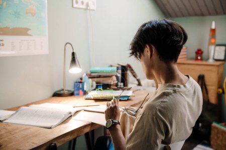 Photo for Serious asian teenage guy studying with book while sitting at desk at home - Royalty Free Image