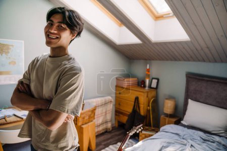 Téléchargez les photos : Jeune homme asiatique portant t-shirt souriant à la caméra tout en se tenant à la maison - en image libre de droit