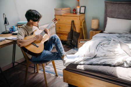 Téléchargez les photos : Jeune homme asiatique en casque jouant de la guitare tout en étant assis sur une chaise à la maison - en image libre de droit