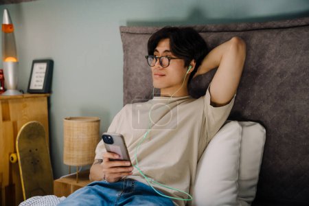 Téléchargez les photos : Jeune homme asiatique dans les écouteurs en utilisant le téléphone mobile tout en se reposant sur le lit à la maison - en image libre de droit