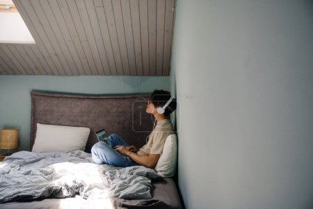 Téléchargez les photos : Jeune homme asiatique dans les écouteurs de travail avec ordinateur portable tout en se reposant sur le lit à la maison - en image libre de droit