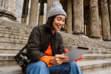 Photo for Beautiful young smiling asian woman with backpack using tablet computer while sitting on stairs at old city street - Royalty Free Image