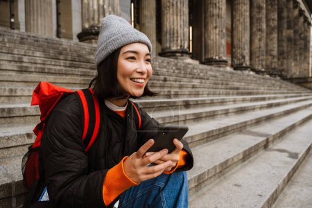 Téléchargez les photos : Belle jeune femme asiatique souriante avec sac à dos en utilisant un smartphone tout en étant assis sur les escaliers à la vieille rue de la ville - en image libre de droit