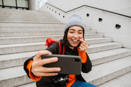 Téléchargez les photos : Beautiful young smiling asian woman in warm clothes taking selfie while sitting on stairs at city street - en image libre de droit