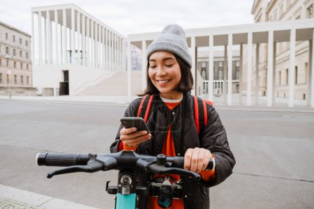 Téléchargez les photos : Young joyful asian woman in warm clothes using mobile phone for electric scooter's rent outdoors at city street - en image libre de droit