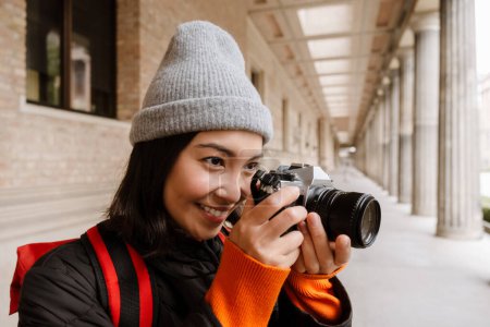 Téléchargez les photos : Beautiful smiling young asian woman tourist with backpack taking pictures with camera during walk through the old city street - en image libre de droit