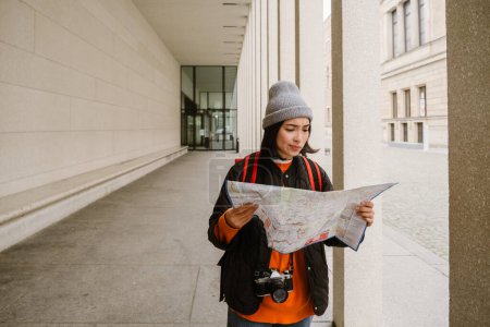 Photo for Lost asian woman in warm clothes examining paper map during walk at city street - Royalty Free Image