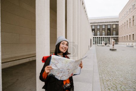 Téléchargez les photos : Jeune femme asiatique joyeuse en vêtements chauds examinant la carte papier pendant la promenade dans la rue de la vieille ville - en image libre de droit