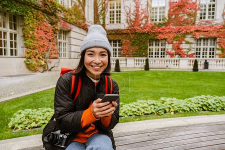 Téléchargez les photos : Cheerful beautiful young asian woman in warm clothes using mobile phone while sitting on bench in old city - en image libre de droit