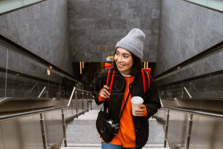 Téléchargez les photos : Beautiful young smiling asian woman in warm clothes drinking takeaway coffee while standing outdoors on stairs - en image libre de droit