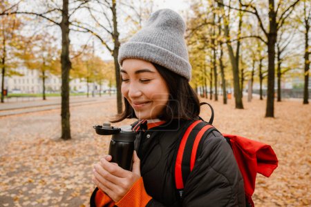 Foto de Hermosa sonriente joven mujer asiática usando ropa de abrigo bebiendo té caliente del termo durante el paseo en el parque de otoño - Imagen libre de derechos