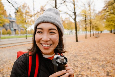 Photo for Beautiful cheerful young asian woman wearing warm clothes drinking hot tea from thermos during walk in autumn park - Royalty Free Image