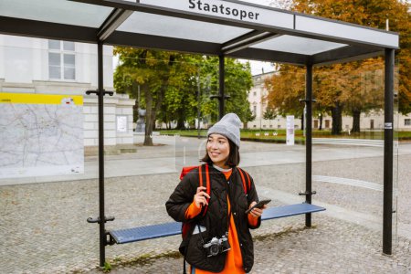 Foto de Joven mujer asiática alegre usando ropa de abrigo usando teléfono inteligente y mirando a un lado mientras está de pie en la estación de autobuses - Imagen libre de derechos
