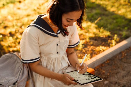 Foto de Young smiling asian woman wearing dress drawing on digital tablet with stylus while sitting in park - Imagen libre de derechos