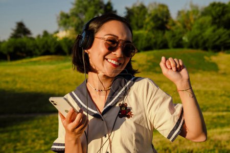 Photo for Smiling young asian girl in sunglasses dancing while listening music with headphones and mobile phone in park - Royalty Free Image