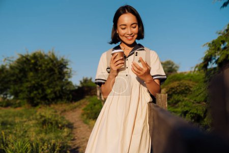 Téléchargez les photos : Young beautiful smiling asian woman with backpack holding coffee and mobile phone while walking outdoors in green park - en image libre de droit