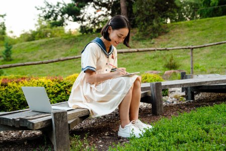 Téléchargez les photos : Young smiling asian woman wearing dress writing down notes while sitting on bench in green park - en image libre de droit