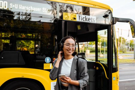 Foto de Joven hermosa sonriente mujer asiática en auriculares sosteniendo teléfono inteligente y mirando a un lado mientras está de pie cerca del autobús al aire libre - Imagen libre de derechos
