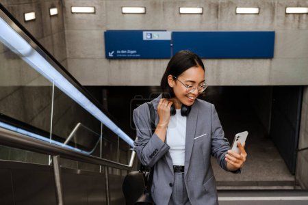 Téléchargez les photos : Jeune femme asiatique joyeuse prenant selfie sur smartphone tout en se tenant debout sur les escaliers de la ville à l'extérieur - en image libre de droit